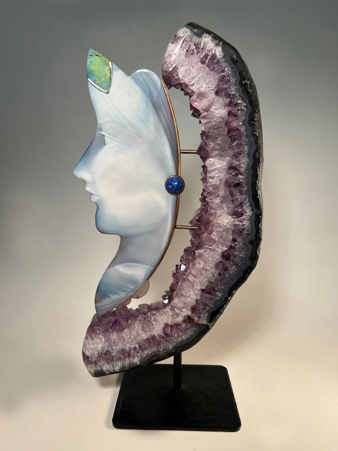 Skulptur mit einem gravierten Gesicht aus hellem Achat, verziert mit einem grünschimmernden Opal auf der Stirn und einem blauen Stein (Lapis) an der Seite, eingebettet in ein Stück einer Amethyst Druse auf schwarzem Ständer