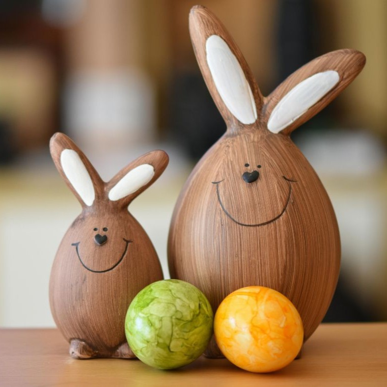 Zwei hölzerne Osterhasenfiguren mit weißen Ohren und lächelnden Gesichtern neben einem grünen und einem gelben Osterei
