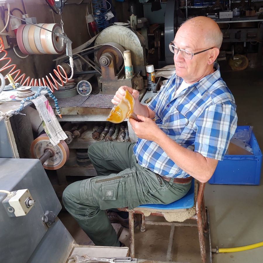 Ein älterer Mann mit Brille und kariertem Hemd sitzt auf einem Stuhl vor einer Schleifmaschine in einer Werkstatt und hält einen gelblichen Edelsteinpokal in der Hand