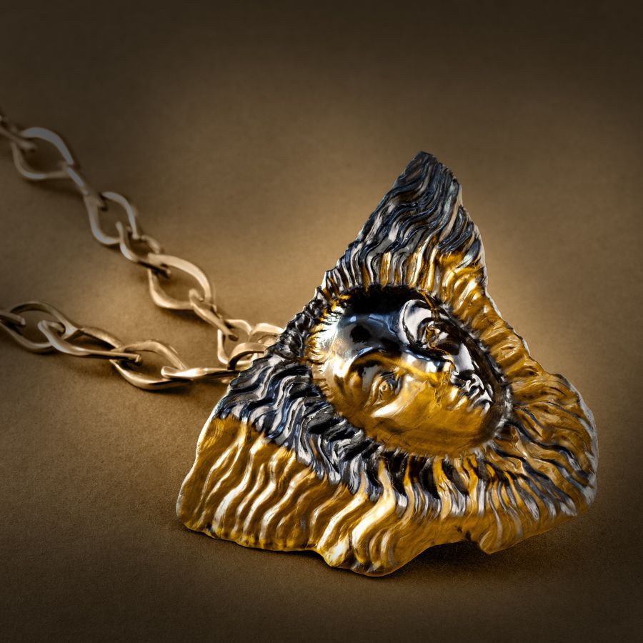 Anhänger in Gelbgold mit graviertem Tigerauge, gestaltet als stilisiertes Gesicht mit wellenförmiger Umrandung an einer Gliederkette